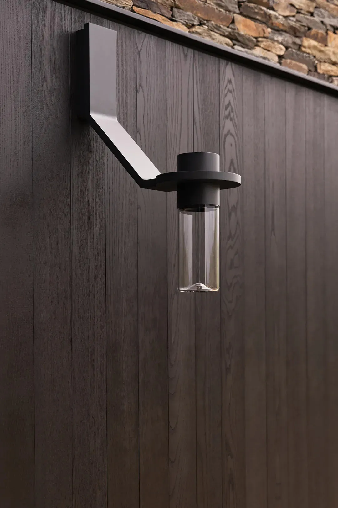 Modern black outdoor wall sconce with cylindrical glass shade mounted on dark vertical wood paneling next to a stone wall.
