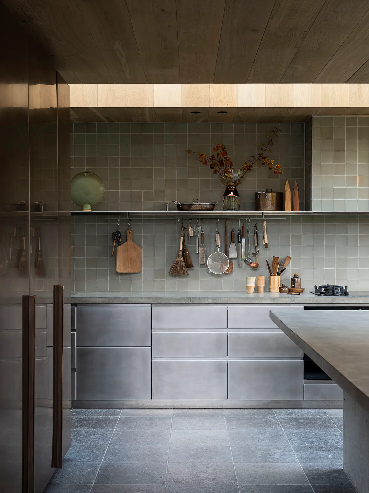 Modern minimalist kitchen featuring concrete cabinetry, grey tiled walls, open shelving with hanging utensils, a concrete island, and warm wood ceilings.