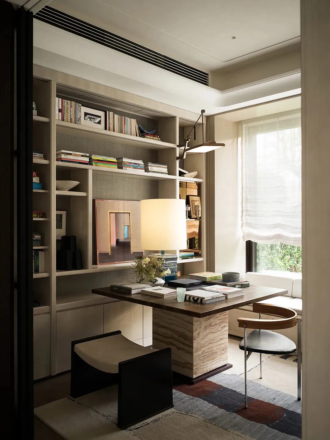 Modern home office interior with a large travertine desk, built-in light wood bookshelves filled with books, a wooden and leather chair, a black bench, and a large window with sheer curtains.