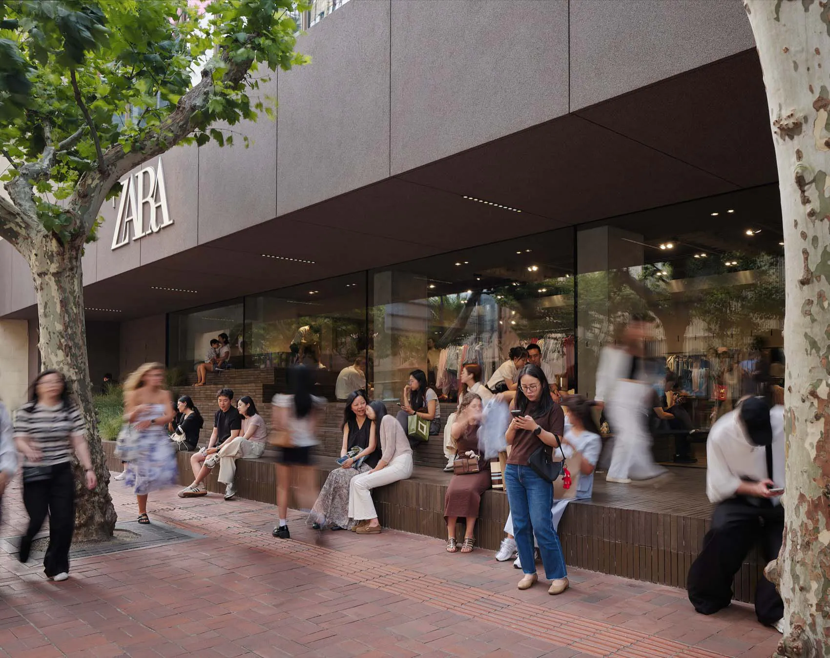 現代Zara店面前熱鬧的都市廣場,大面玻璃窗、洞石階梯作為座椅,人們在陽光下的大樹旁休憩。