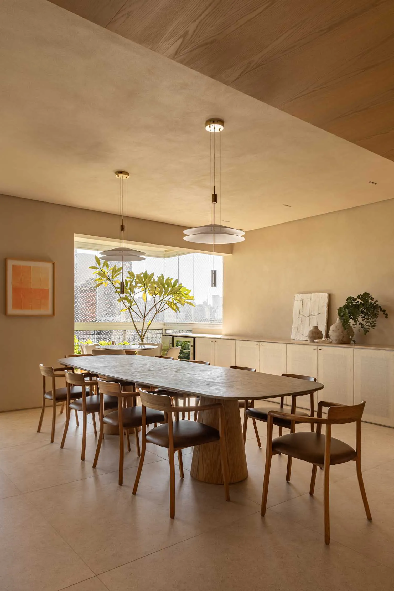 Modern minimalist dining room featuring a large oval terrazzo dining table with a fluted wooden pedestal base, surrounded by eight wooden dining chairs with brown leather seats, under two large white pendant lights, with a large window overlooking a city view and a potted plant in the background.
