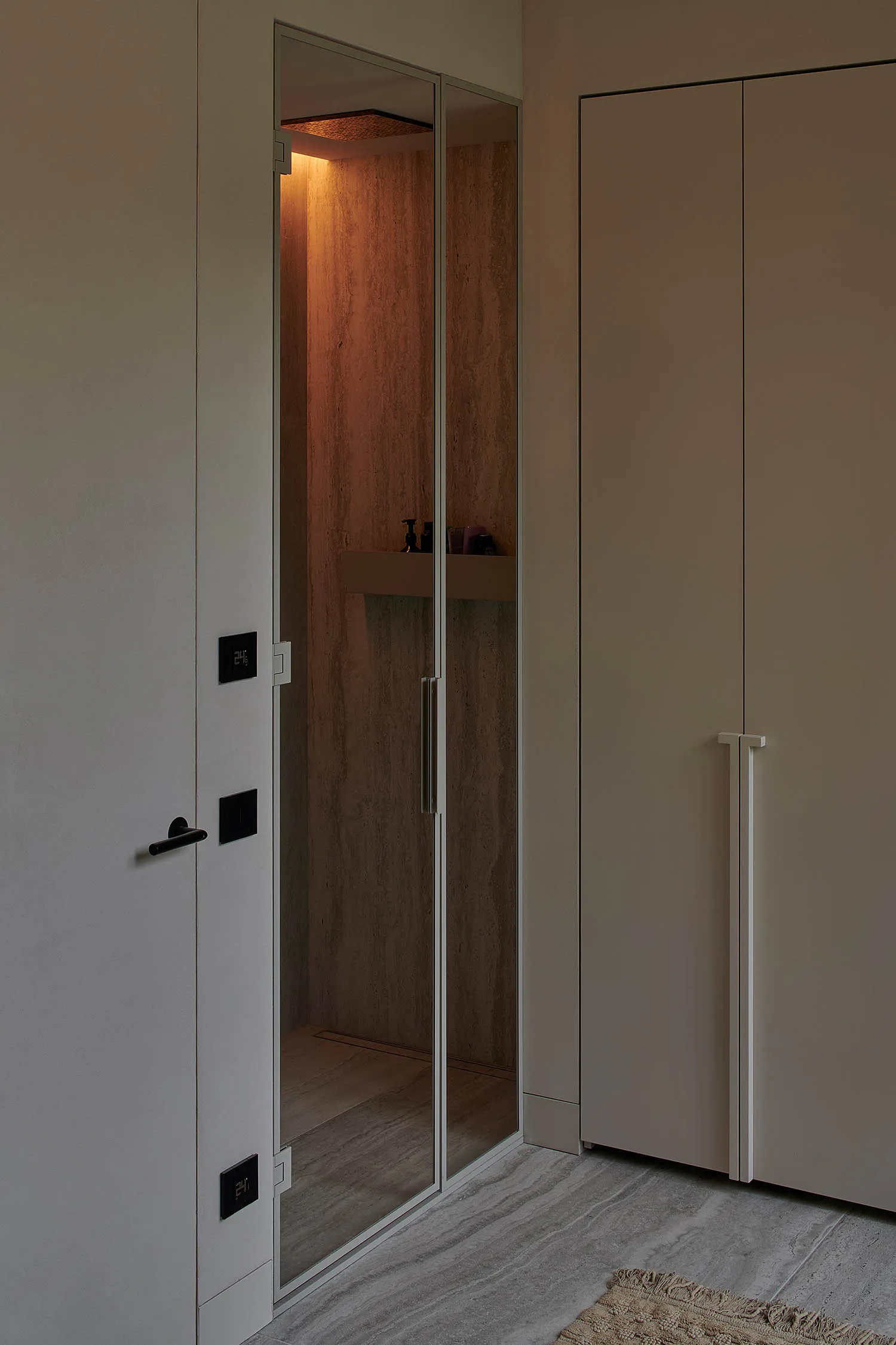 Modern minimalist walk-in steam shower with travertine stone walls, warm ambient lighting from above, glass door, built-in shelf with toiletries, and adjacent beige cabinetry in a neutral-toned bathroom.