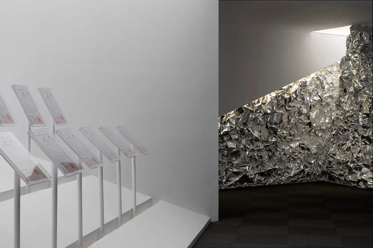 Modern art gallery interior featuring a large wall covered in crumpled reflective silver foil next to a row of white acrylic sign stands on a white platform.
