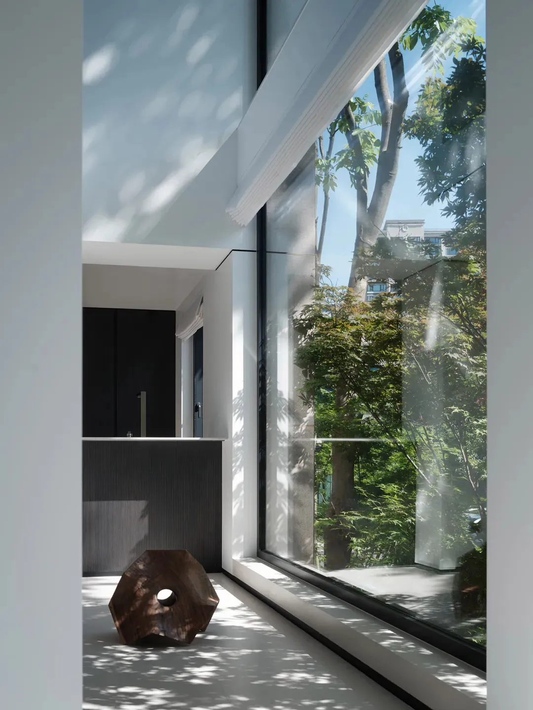 A bright, minimalist modern living room interior with white walls, large floor-to-ceiling windows overlooking green trees, a dark wood counter in the background, and a distinctive dark wooden sculptural stool on the floor in the foreground.