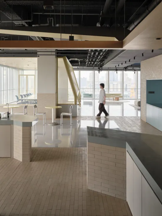 Modern open-plan office kitchen and lounge area with white brick counters, pale wood herringbone floor, round tables, and large windows overlooking a river and city skyline. A man in a white shirt walks across the reflective floor.