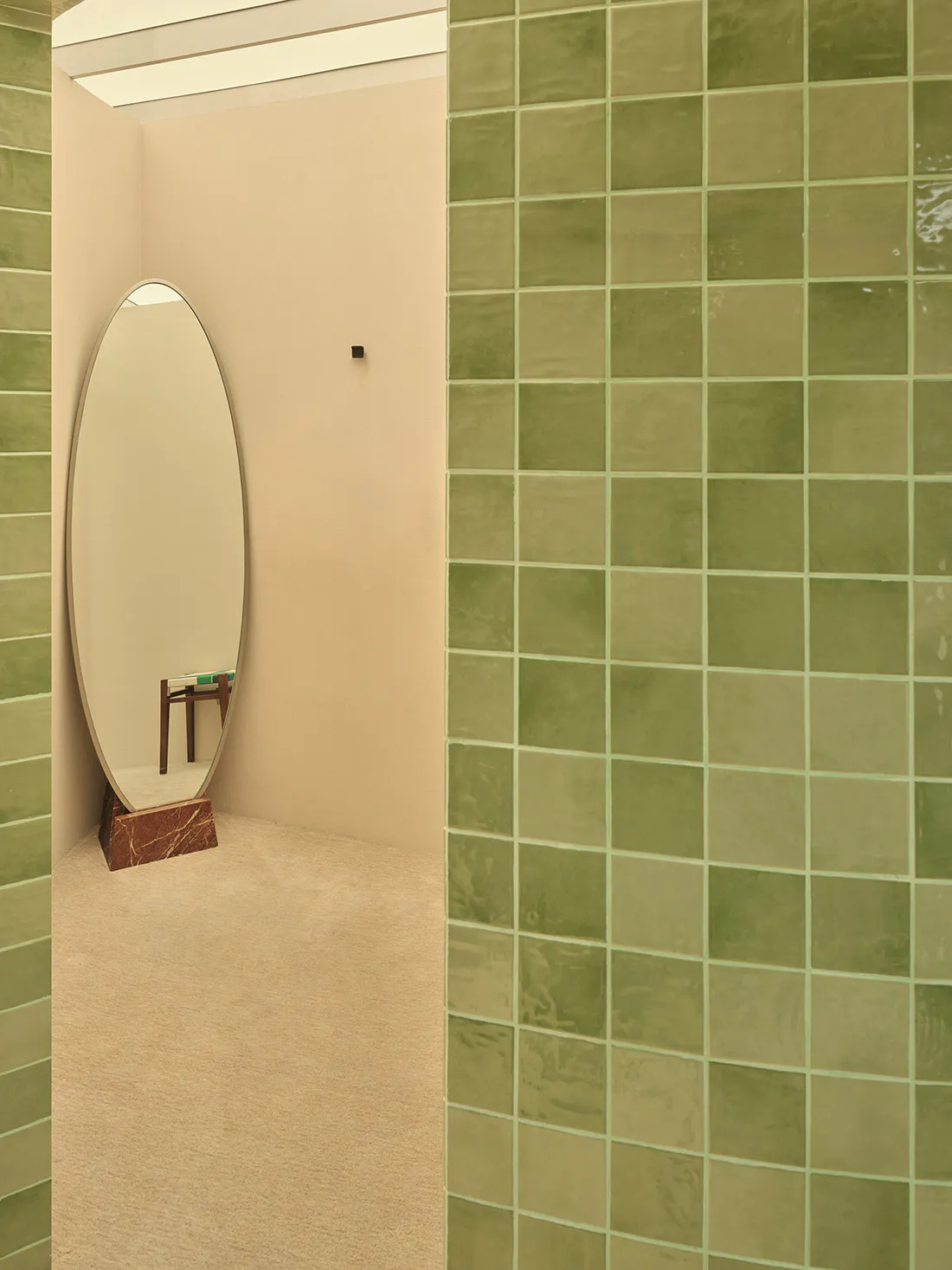Modern entryway corner featuring glossy sage green ceramic tile walls, a large oval floor mirror with a red marble base, and beige carpeted floor.