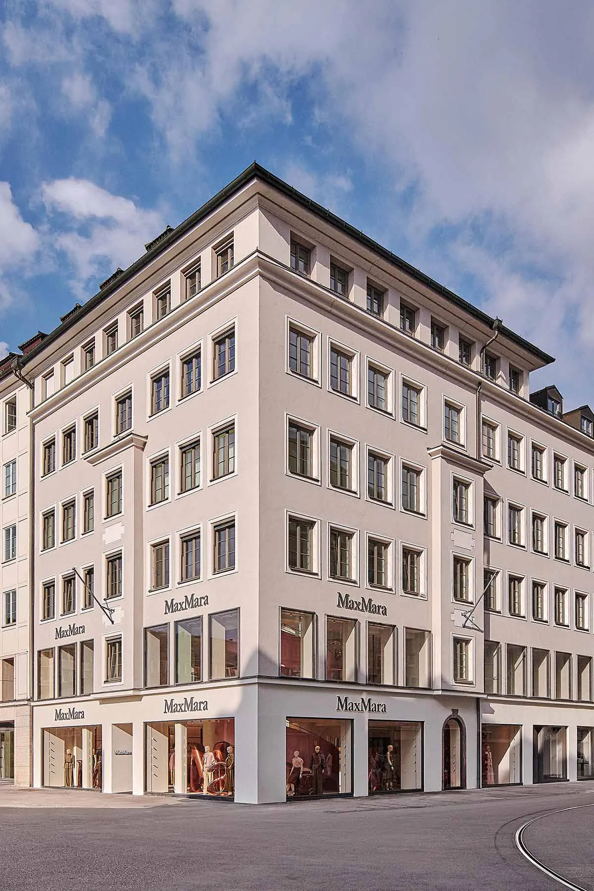 Exterior view of a multi-story white MaxMara boutique building on a street corner with large ground-floor display windows and mannequins.
