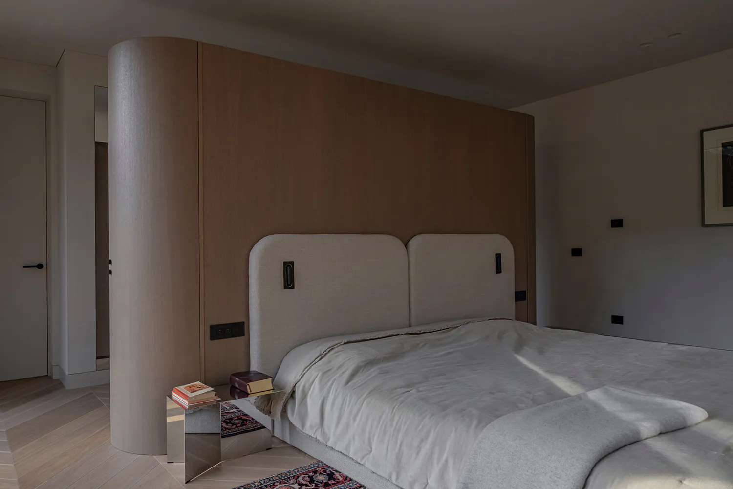 Modern minimalist bedroom with a large curved light-wood wall unit serving as a headboard, two upholstered beige headboards, a metallic mirrored nightstand with stacked books, and a patterned Persian-style rug on a light wood floor.