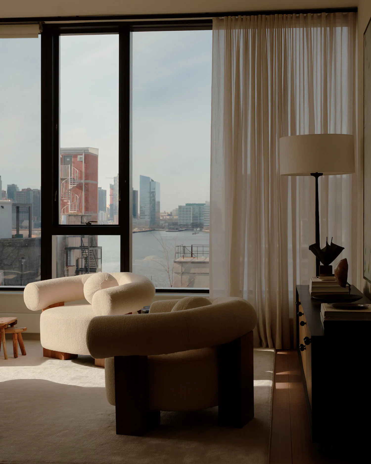 Modern living room interior featuring two cream boucle armchairs with dark wood bases, a black console table with a tall floor lamp, sheer curtains, and a large window revealing a panoramic view of the New York City waterfront with brick buildings and the river.