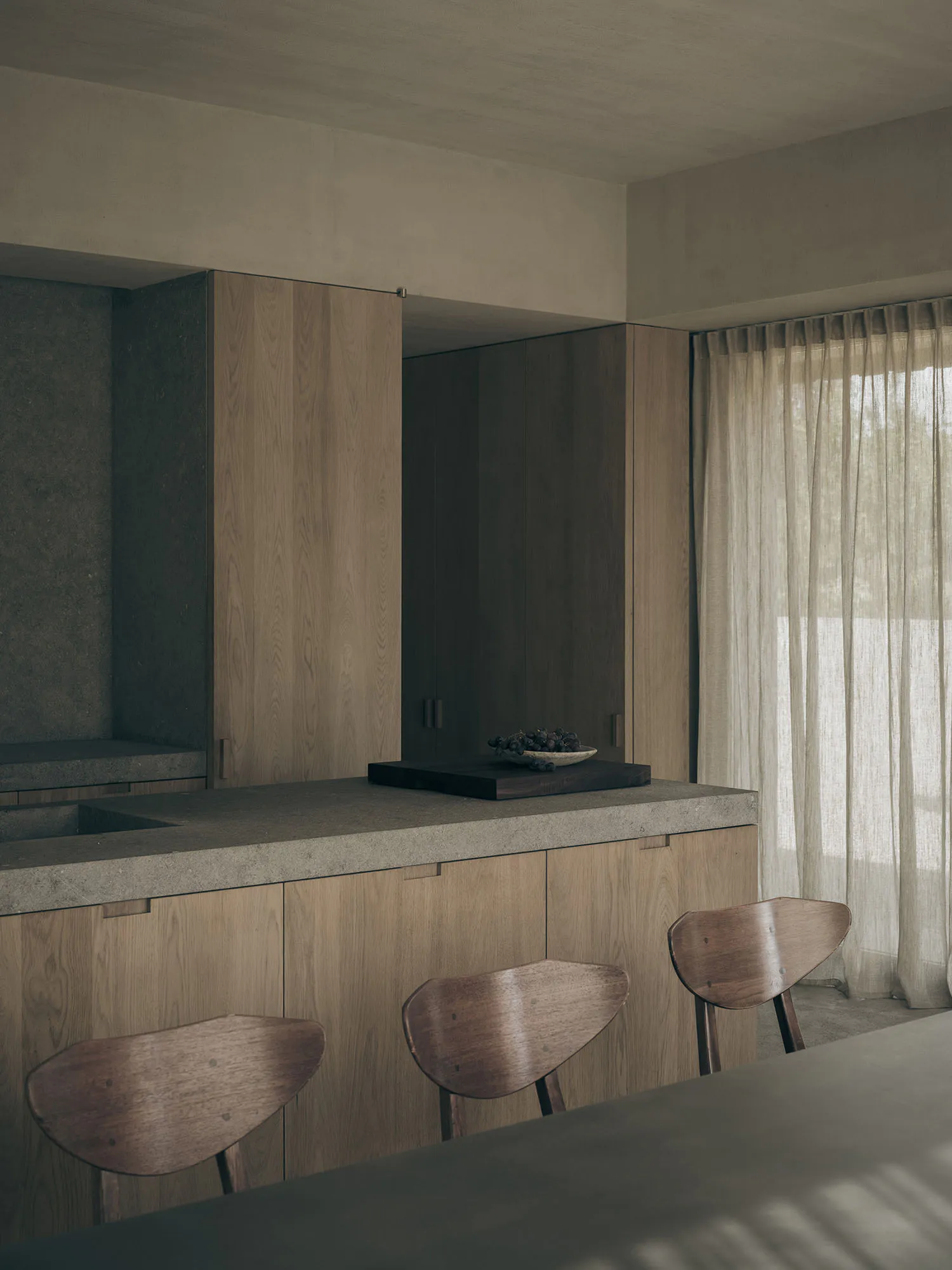 Modern minimalist kitchen featuring a concrete countertop island, light oak cabinetry, wooden butterfly chairs, and a dark concrete accent wall with a bowl of fruit on a wooden cutting board.