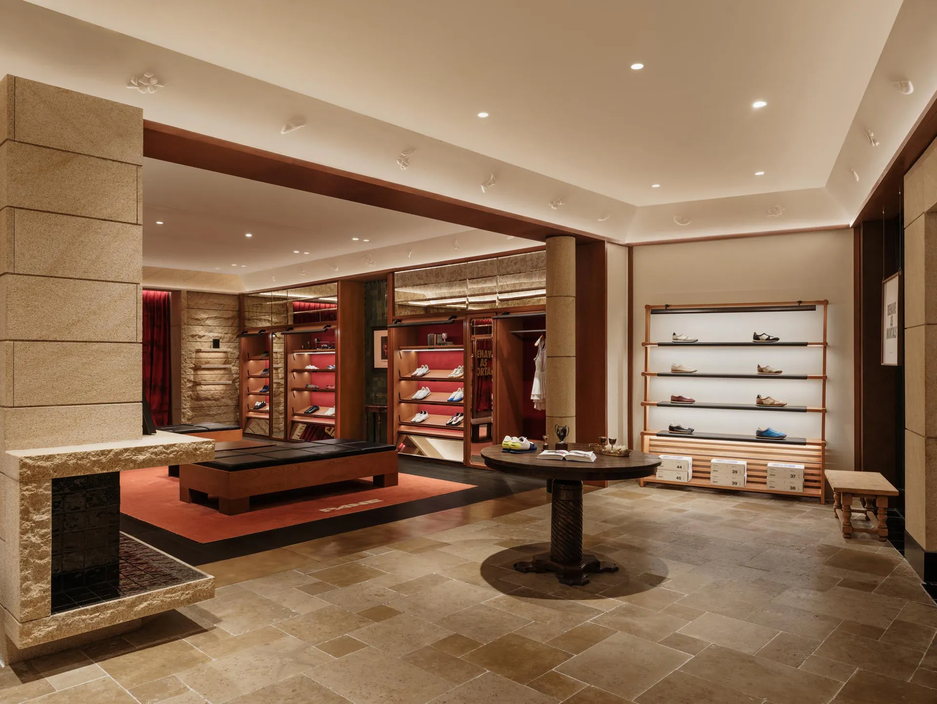 Interior view of a high-end shoe boutique with a large travertine fireplace on the left, built-in wooden display shelves with red interiors holding various shoes, a central round dark wood table on a circular rug, and additional floating shelves on the right wall displaying more footwear.