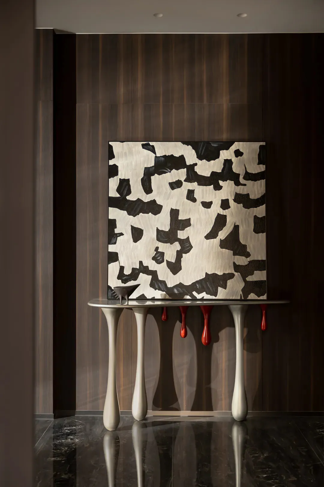 Modern interior scene featuring a sculptural off-white console table with four curved legs, a glossy black marble floor, dark wood paneling, and a large textured black and white abstract painting hanging above it. Red drip-like sculptures hang from the table surface.