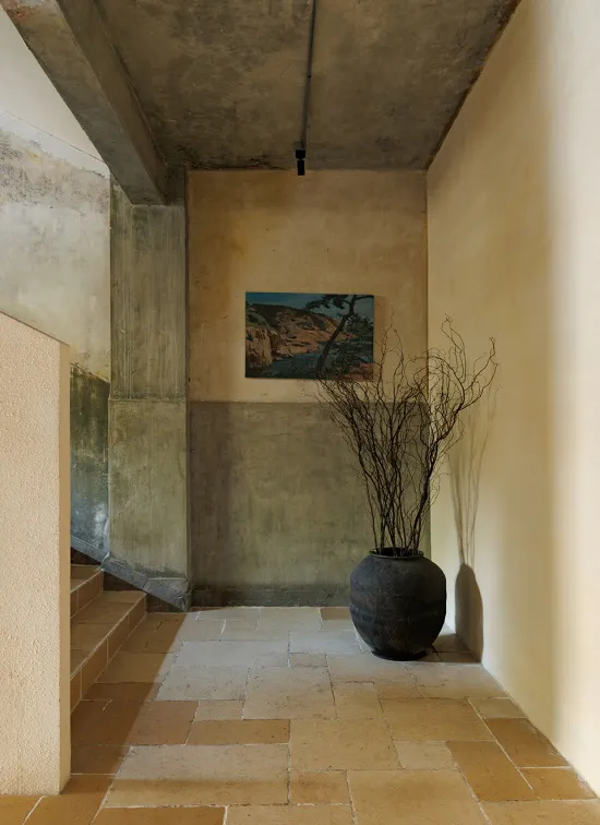 Narrow architectural entryway with raw concrete walls, terracotta stone tile floor, a large dark ceramic vase containing dried branches, and a small landscape painting on the back wall.