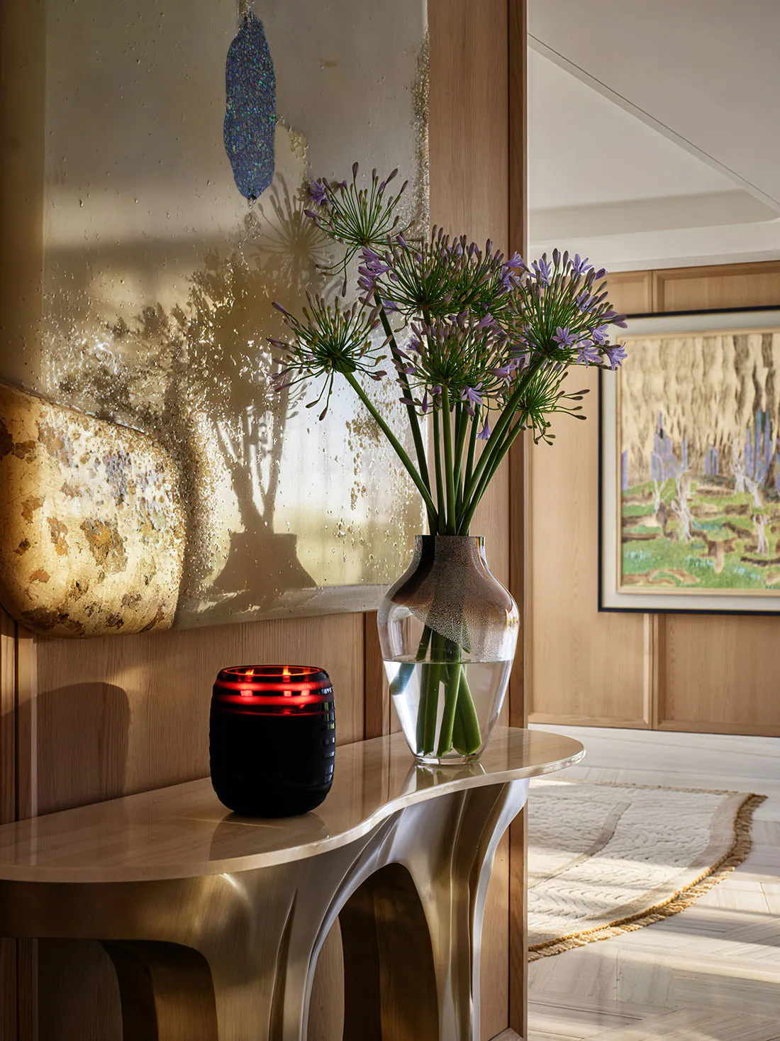 Curved brushed-brass console table displaying a glass vase of purple agapanthus flowers, a glowing red candle holder, and an abstract textured painting in a sunlit entryway.