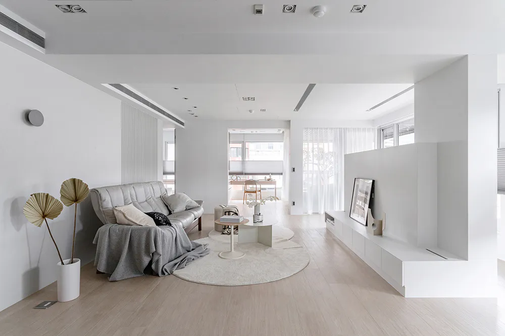 Modern minimalist living room with white walls, light wood flooring, grey leather sofa, round white rug, and large dried palm leaves in a white vase.
