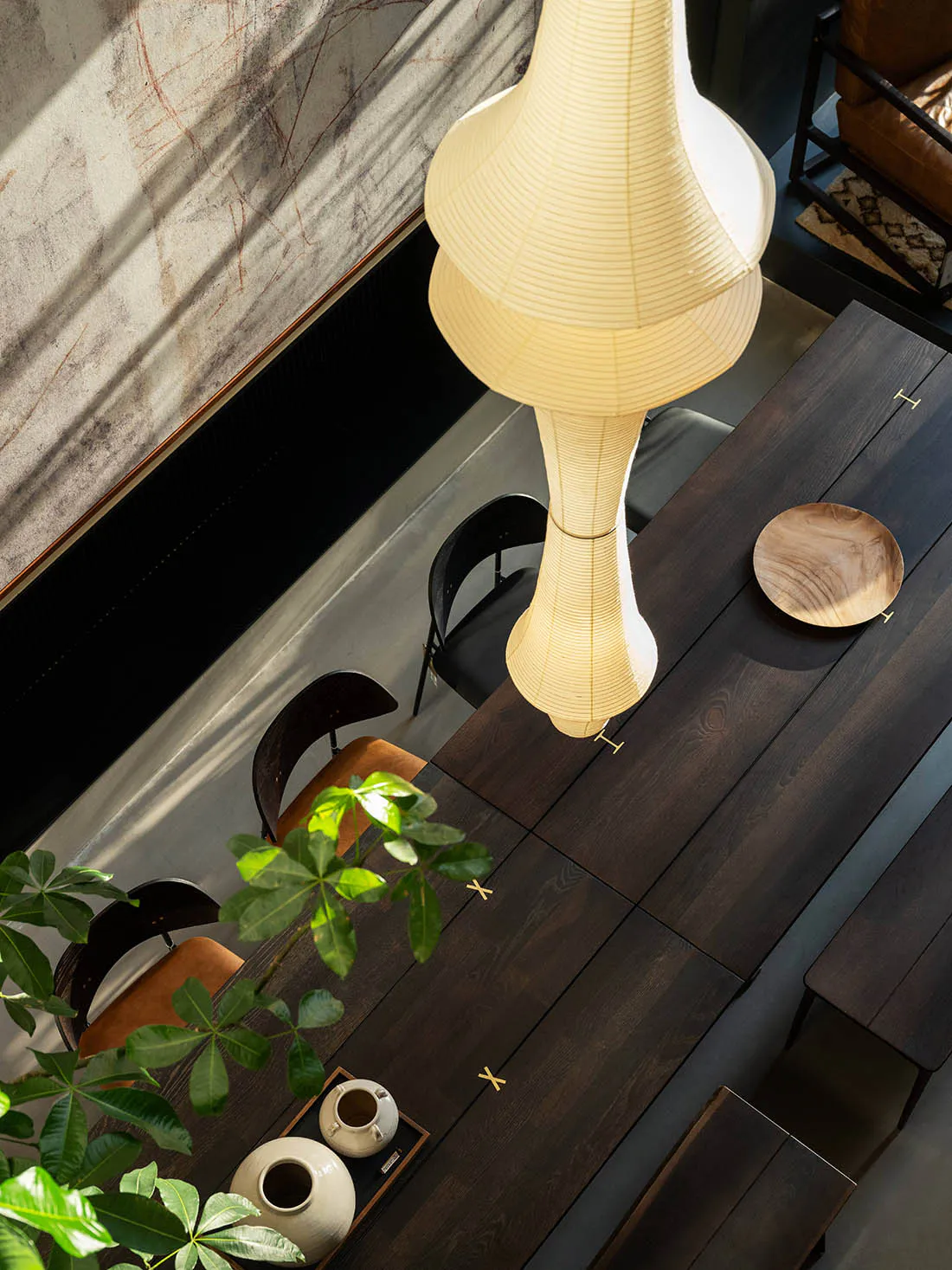 High-angle view of a modern dining room with a dark walnut wood table, black and brown leather chairs, a large sculptural paper pendant lamp, and a potted plant in the foreground.