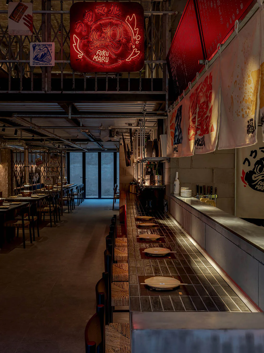 Interior view of a Japanese izakaya featuring a long tiled sushi counter, wooden stools, exposed concrete walls, hanging noren banners, and a prominent glowing red neon Oni mask sign.