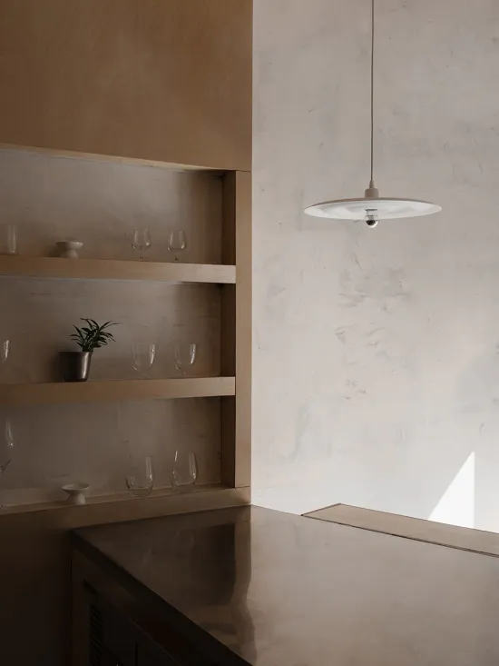 Modern minimalist kitchen corner featuring raw concrete walls and counter, light oak open shelving displaying glassware and a small potted plant, with a white disc pendant light hanging above.