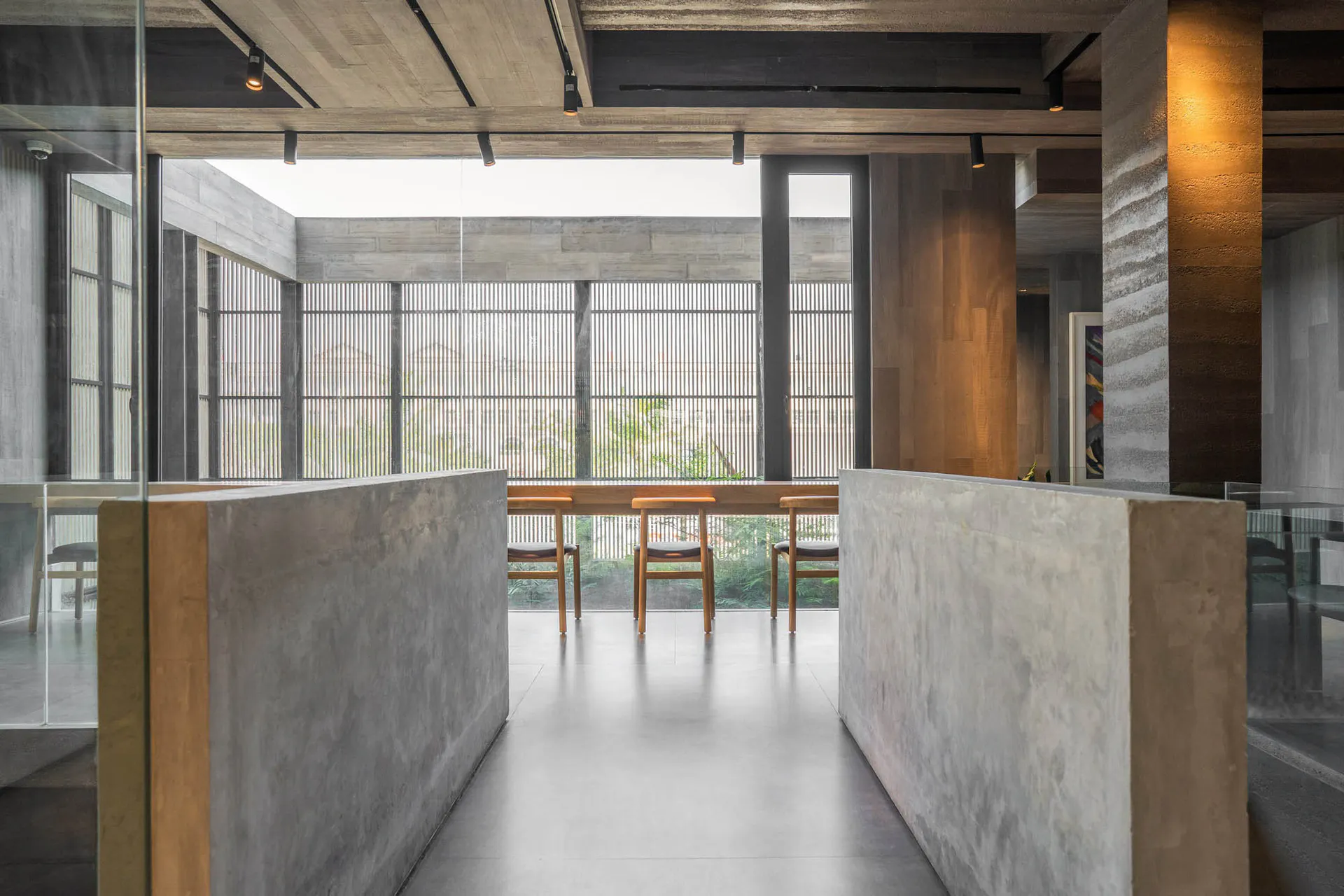 現代極簡用餐區,設有大型清水混凝土隔板、木製餐椅、長木桌,以及可眺望綠意的大型垂直格柵窗戶。