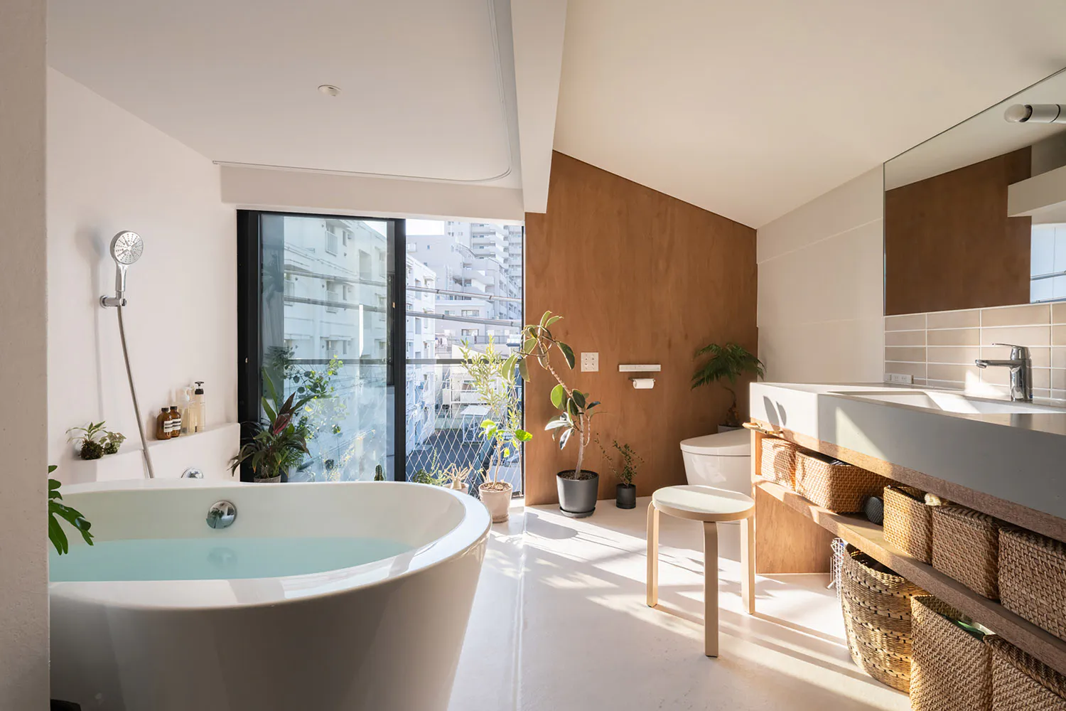 Modern minimalist bathroom interior with white freestanding bathtub filled with water, wooden accent wall, multiple potted plants, wall-mounted faucet, woven storage baskets, and large glass doors opening to a city balcony.
