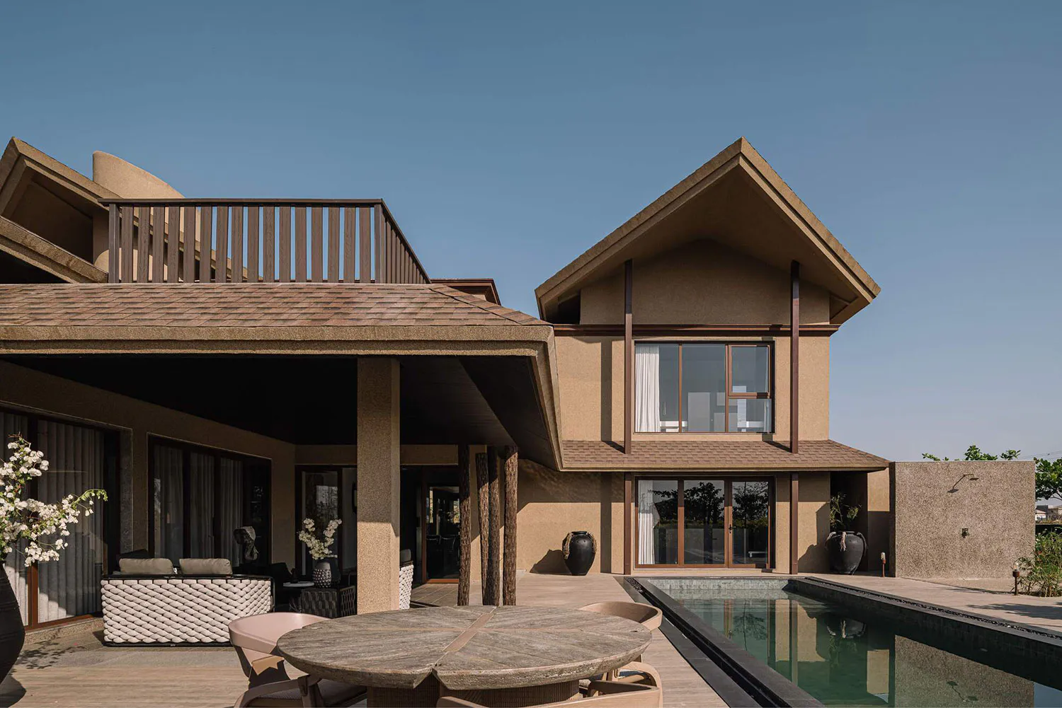 Modern luxury villa exterior featuring a travertine outdoor patio with round wooden dining table, woven lounge furniture, rectangular infinity pool, and two-story beige stucco house with wooden beams and large windows.