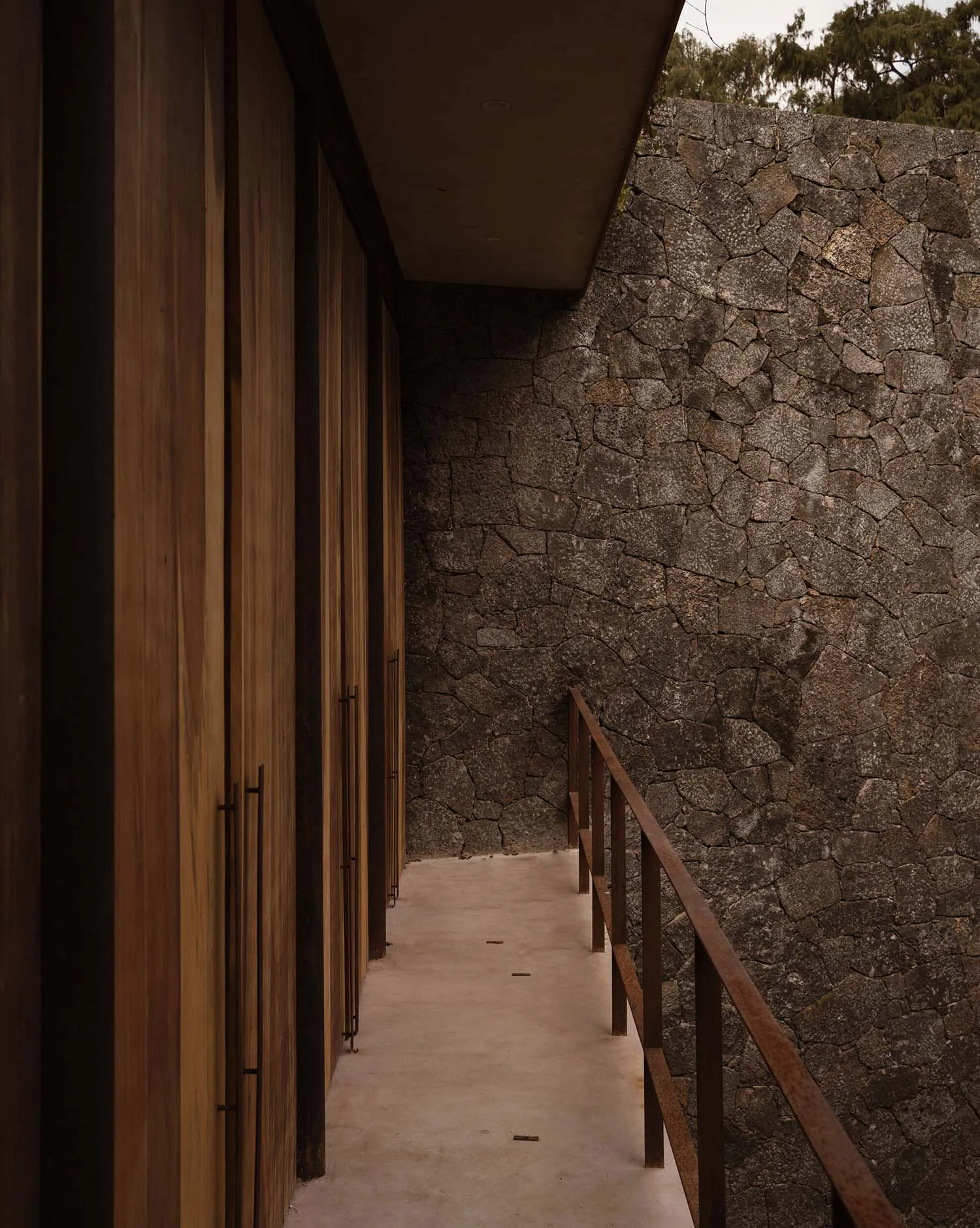 Narrow outdoor corridor featuring vertical teak wood cladding on the left and a rough stone wall on the right, with a metal handrail overlooking the edge.
