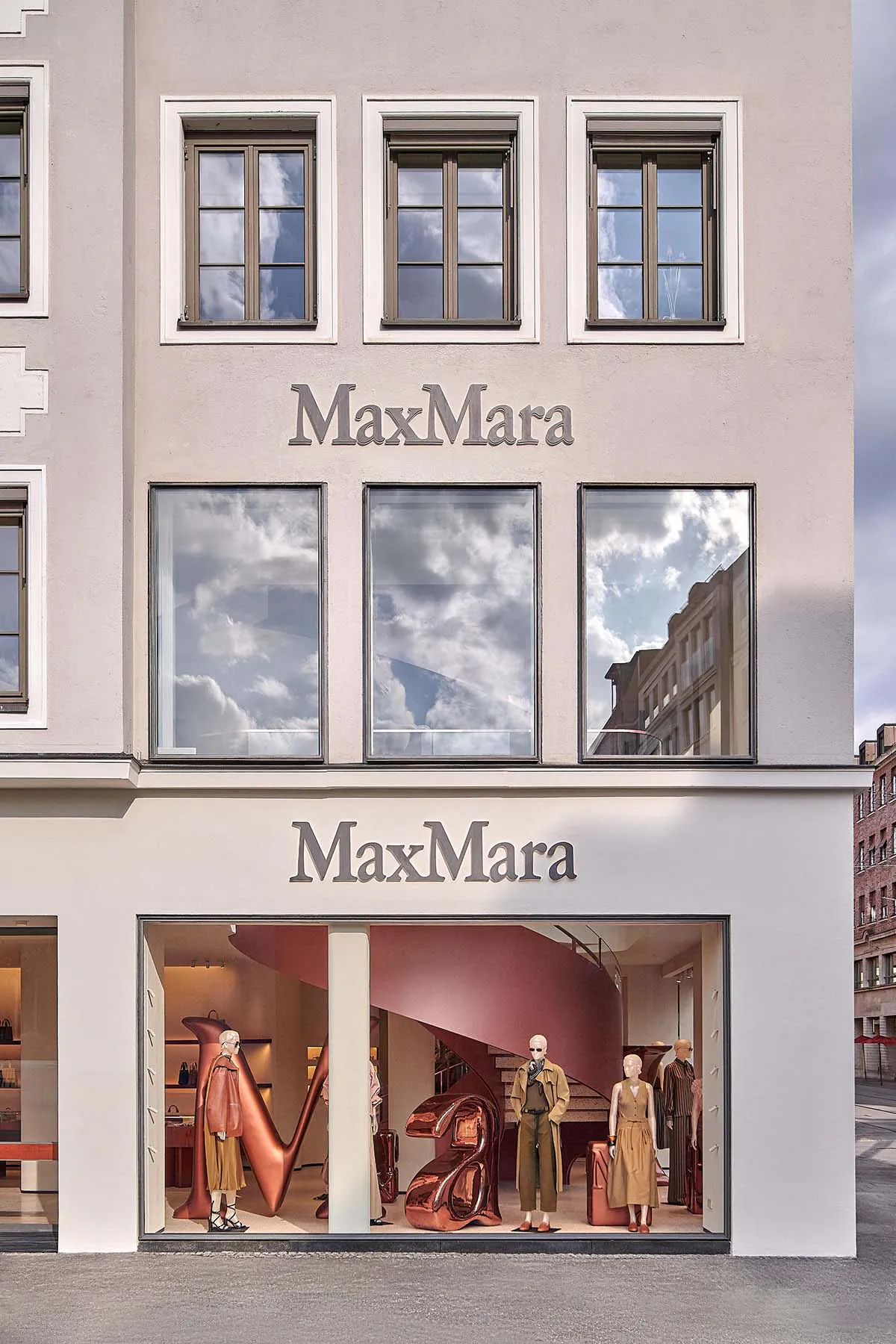 Exterior and interior view of a MaxMara luxury fashion store with large copper sculptures, mannequins in earth-toned outfits, and a pink spiral staircase visible through the glass windows.