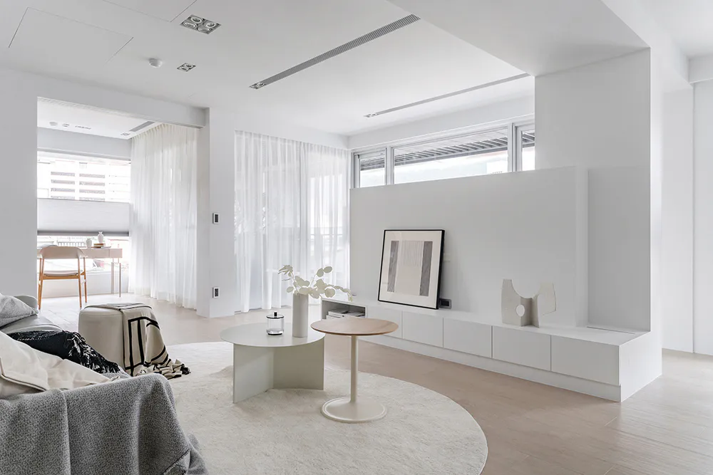 Modern minimalist living room with white walls, light wood flooring, a round white rug, low white oak media console, round coffee tables, and large windows with sheer curtains.