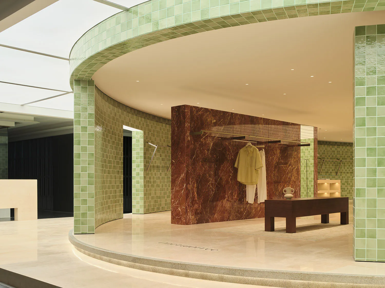 Modern boutique interior featuring curved walls covered in light green tiles, a dramatic red marble display wall with clothing rack, dark wood bench, and polished stone floor under a skylight.