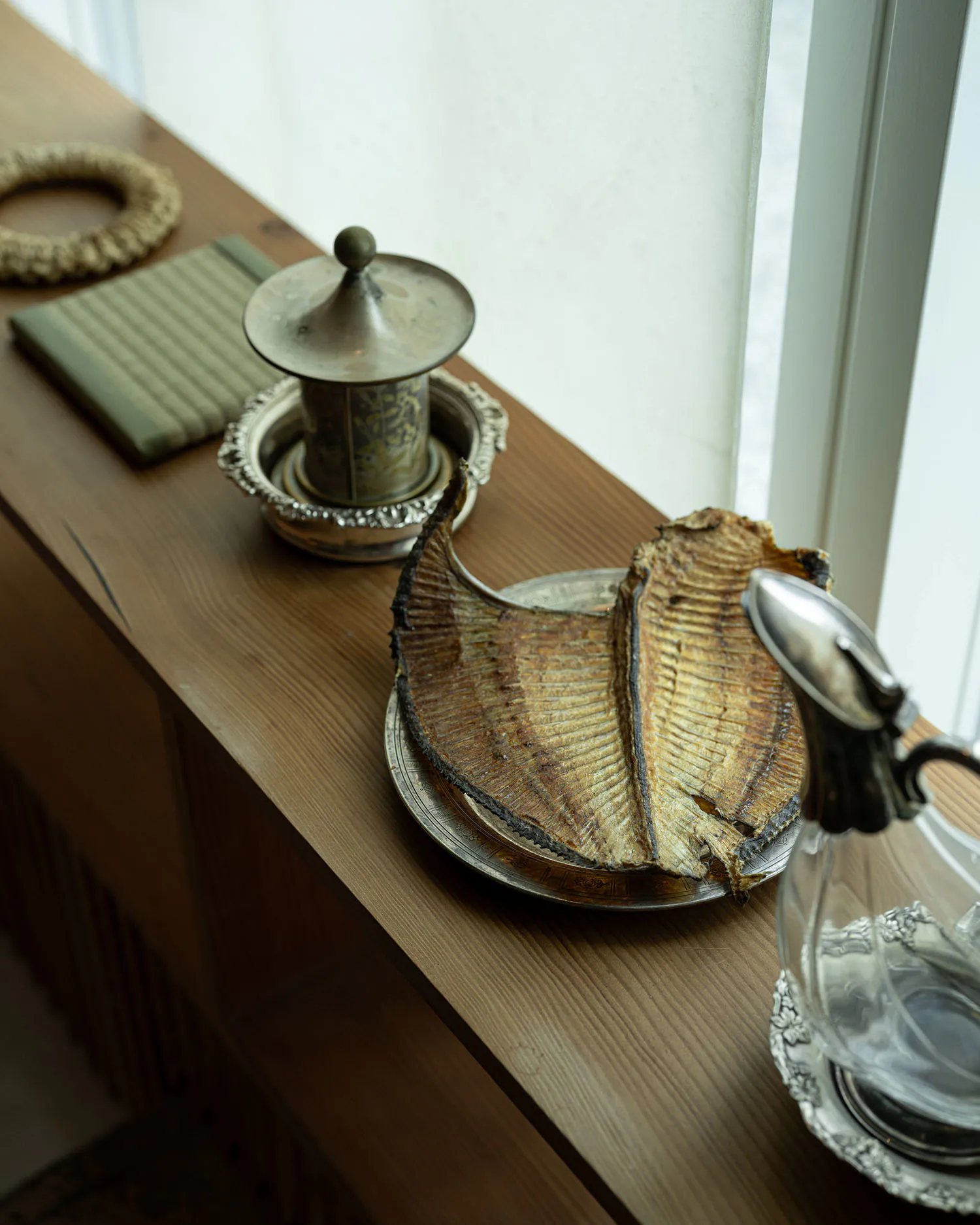 Still life composition on a wooden windowsill with two pieces of dried fish on an ornate silver plate, an antique lidded silver container, a green striped book, a woven ring, and a glass pitcher with silver lid and tray.