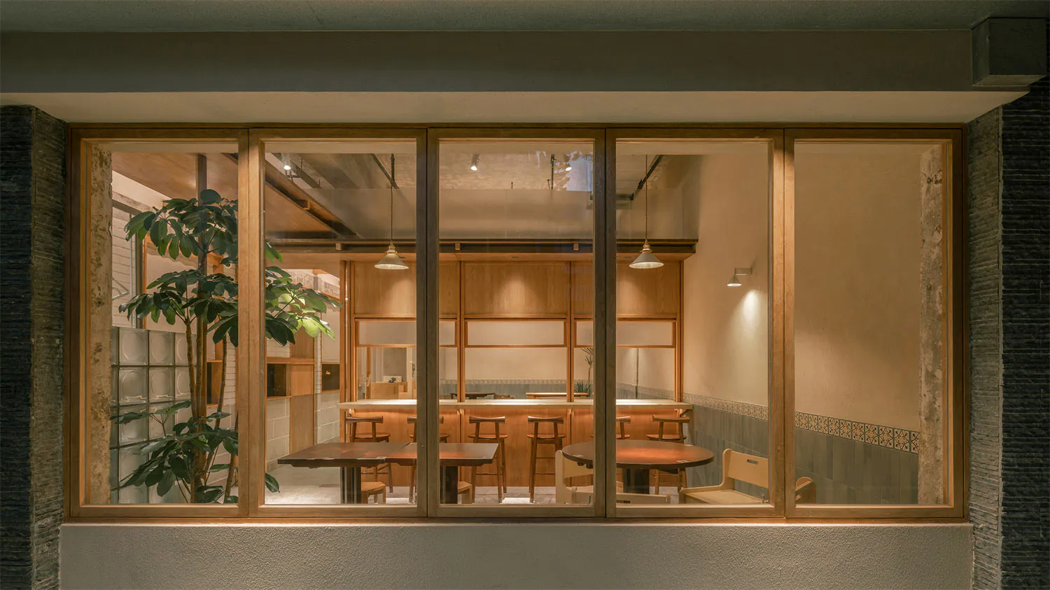 Exterior night view through large wooden-framed glass windows into a warmly lit Japanese restaurant featuring oak furniture, wooden counters, pendant lights, and indoor plants.
