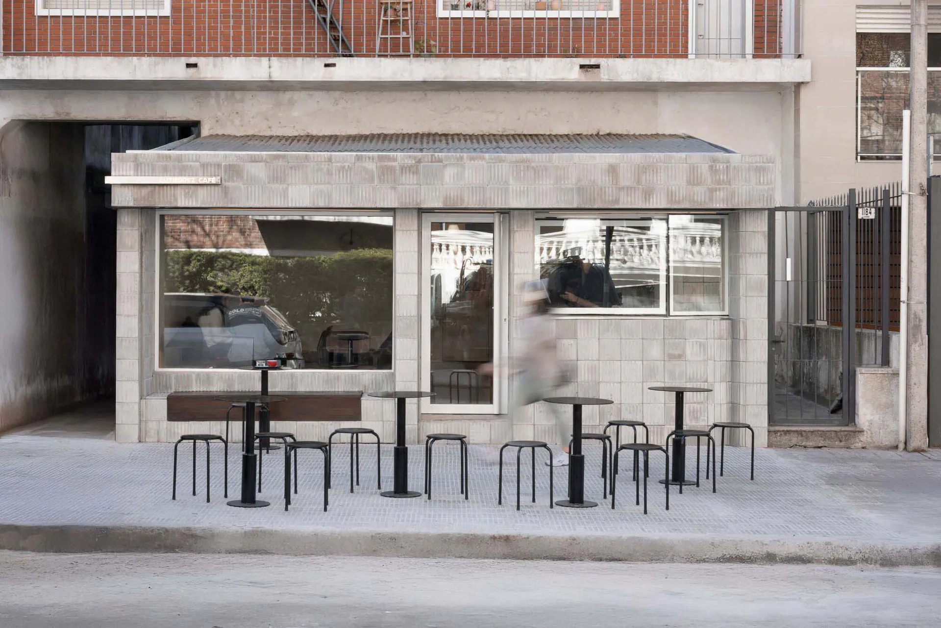 Minimalist concrete cafe storefront with large glass windows, black round tables and stools on a light gray tiled patio, red brick building in background.