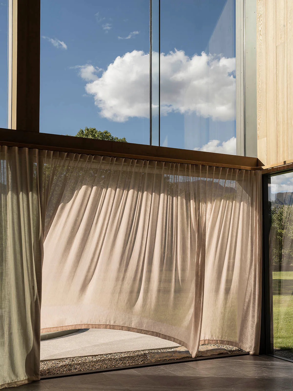 Sheer beige linen curtain billowing in the wind inside a modern room with large glass windows overlooking a garden and mountains.