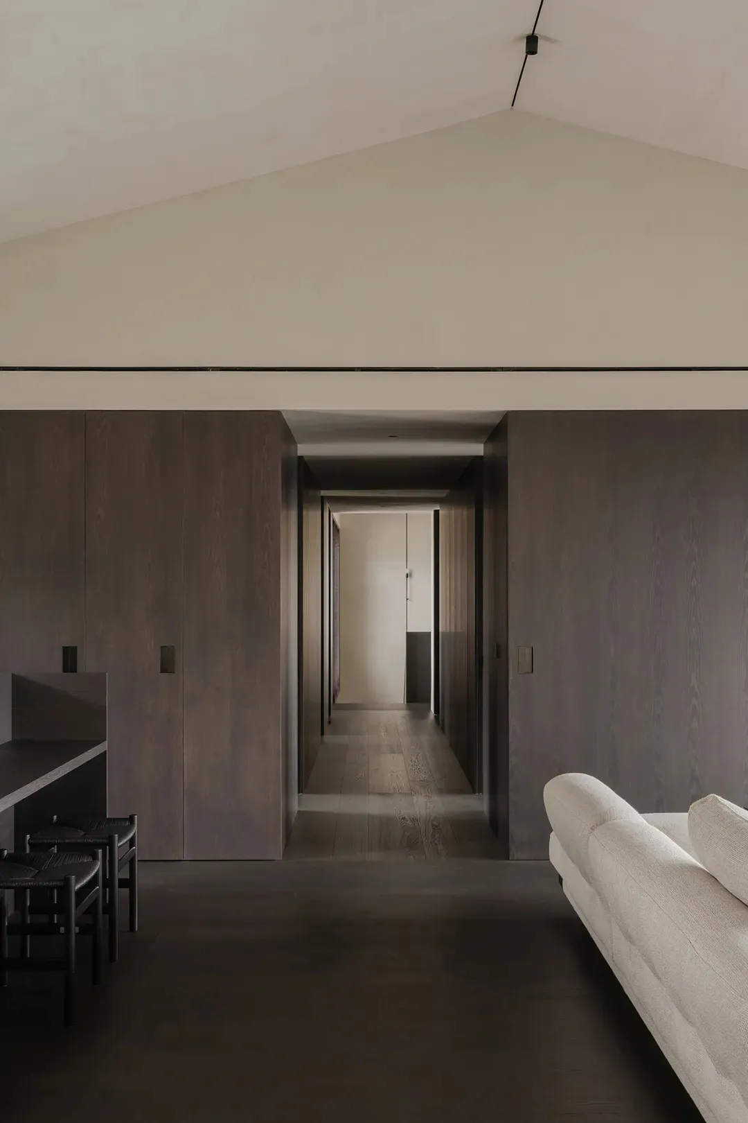 Modern minimalist hallway featuring dark wood cabinetry, a long corridor with light wood flooring, a white fabric sofa in the foreground, and a high vaulted ceiling with a single pendant light.