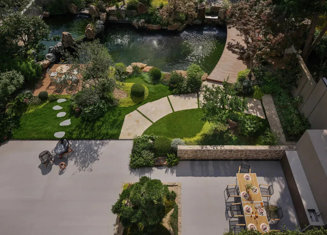 Aerial photograph of a luxurious rooftop garden featuring a large koi pond with waterfall, wooden boardwalk, stone pathways, manicured lawns, and two separate outdoor dining and seating areas with modern furniture.