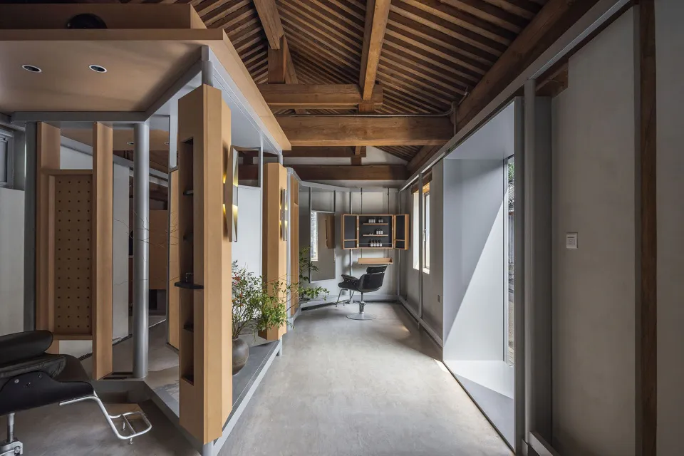 Modern minimalist hair salon interior featuring exposed wooden ceiling beams, polished concrete floors, light wood cabinetry, and natural daylight streaming through large windows.