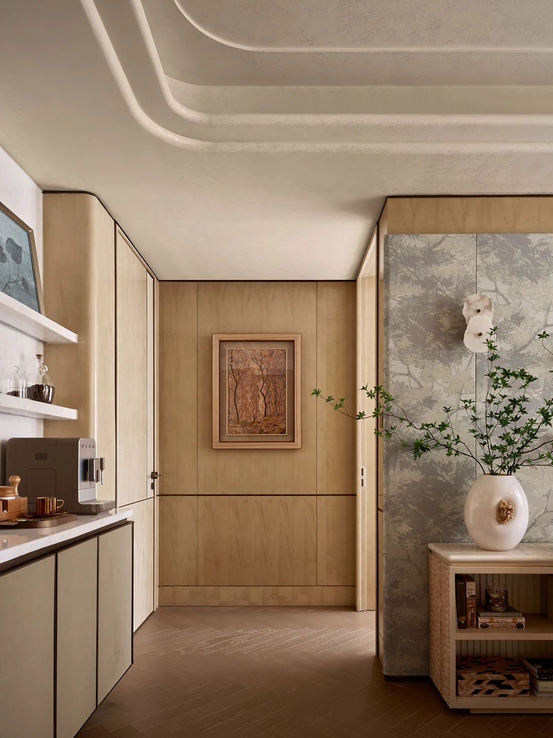 Modern minimalist entryway with light oak built-in cabinetry, marble countertops, sculptural wavy ceiling, framed forest artwork, and a console table with botanical wallpaper and a large white vase with green branches.