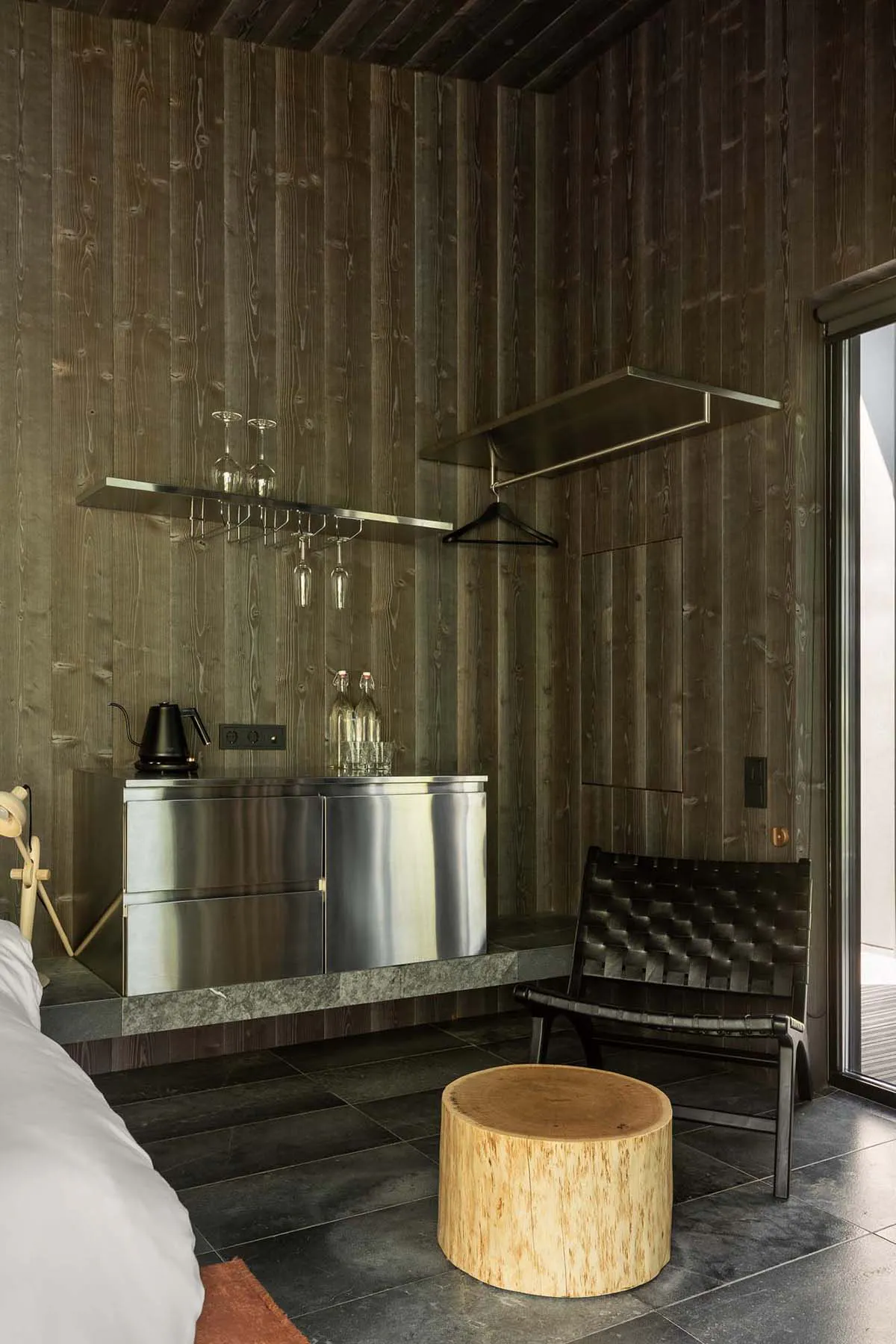 Modern hotel room corner with dark wood walls, stainless steel minibar, floating glass shelves with wine glasses, black leather woven chair, and a tree stump side table.