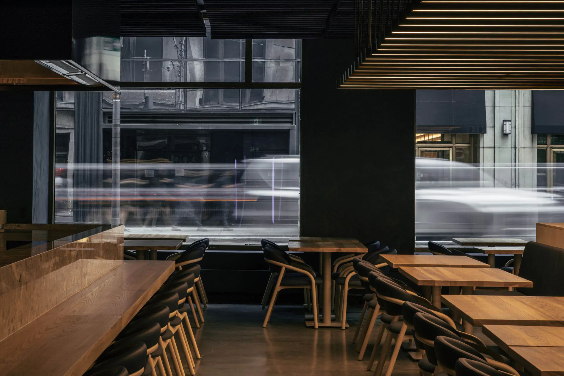 Modern restaurant interior featuring dark oak tables and chairs, black upholstery, dramatic linear ceiling lights, and large windows showing motion-blurred traffic outside.