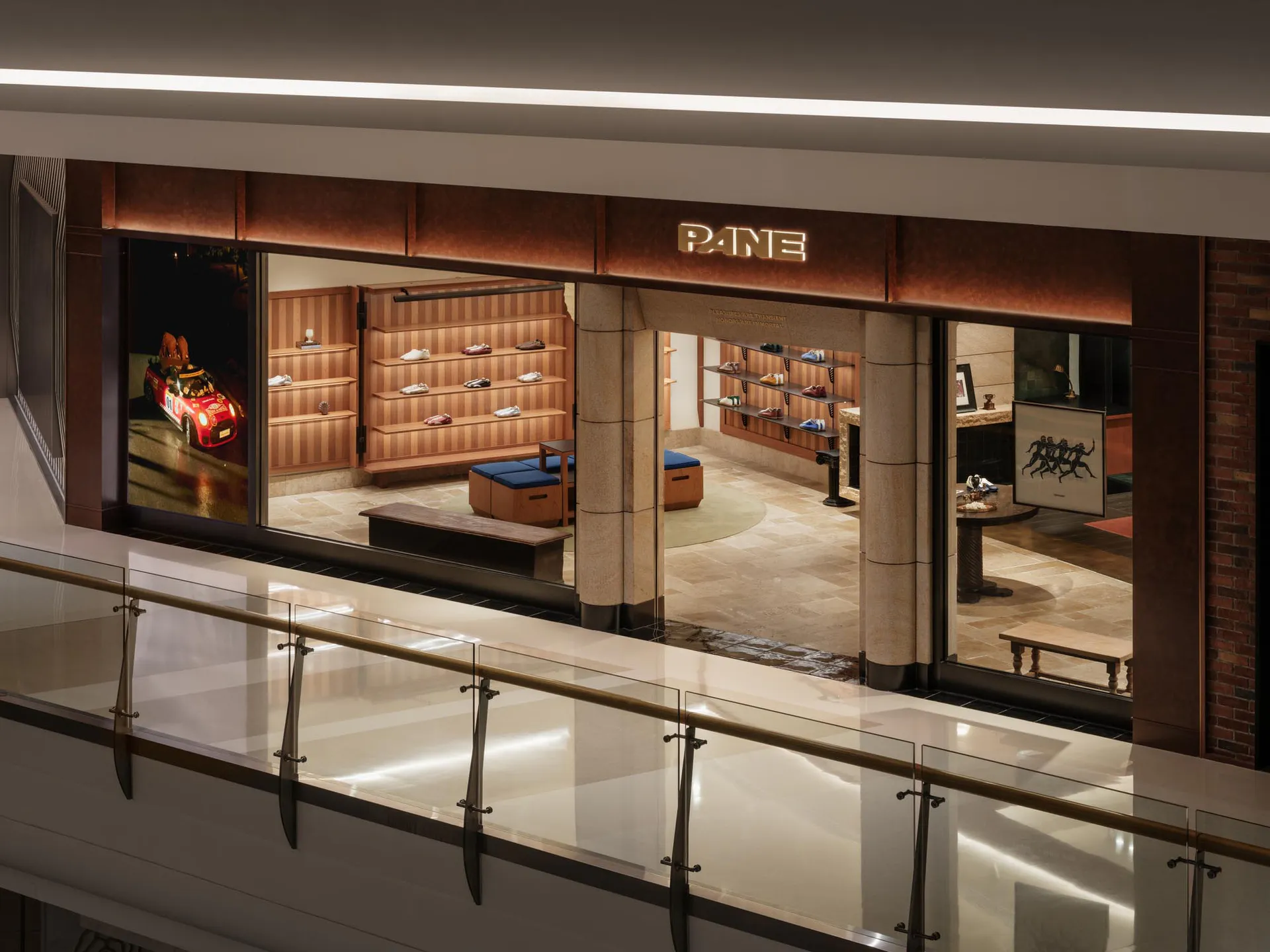 High-angle view of a luxury shoe store named PANE with warm wooden shelving, blue ottomans, stone flooring, and a large red race car photograph on the left wall.