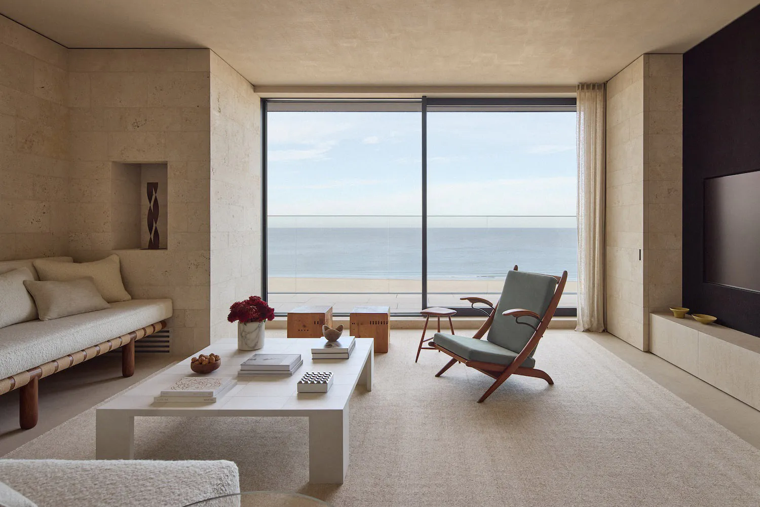 Modern minimalist living room with travertine walls, a built-in daybed, white coffee table, and a wooden lounge chair facing a large glass window overlooking the ocean and beach.