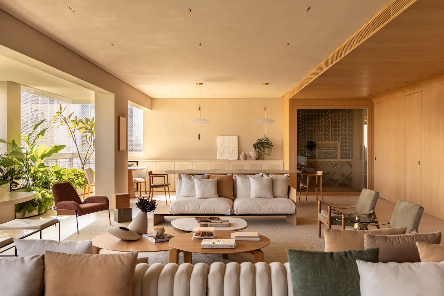 Spacious modern living room featuring a large white sectional sofa, round wooden coffee tables, oak wall paneling, pendant lights, and abundant indoor plants with city views through large windows.