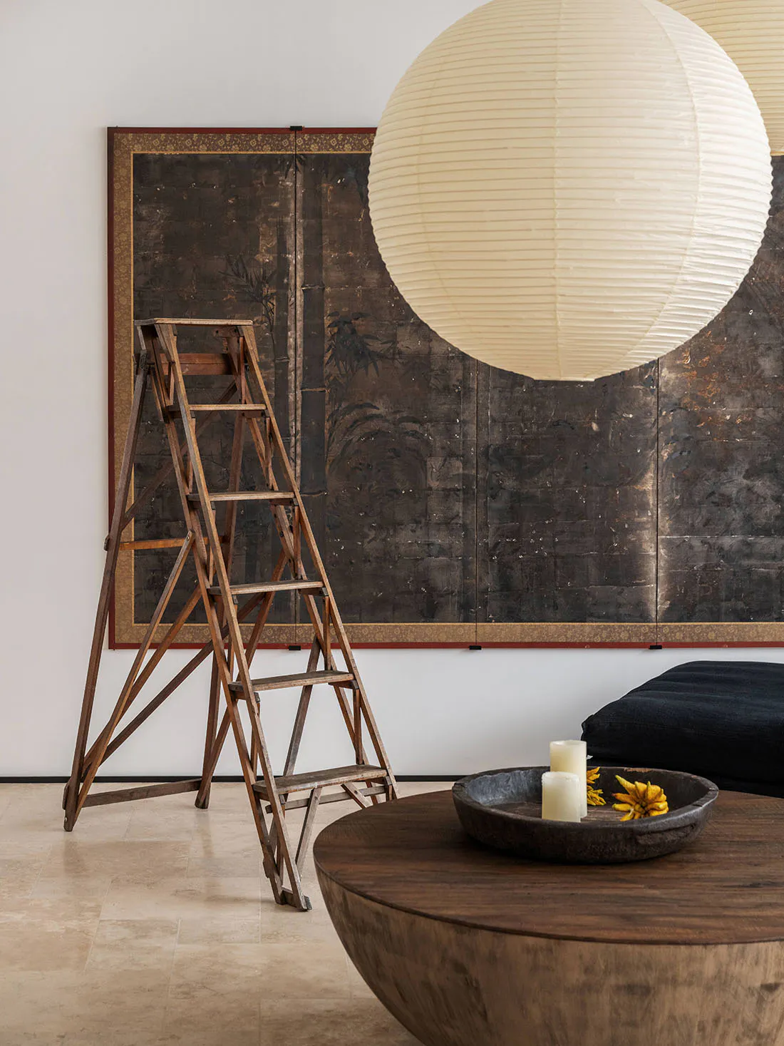 Interior scene featuring a tall wooden A-frame ladder, a large dark antique Japanese byobu screen with gold detailing and nature motifs, two large white paper lanterns, a low round dark wood coffee table, and a black floor cushion.