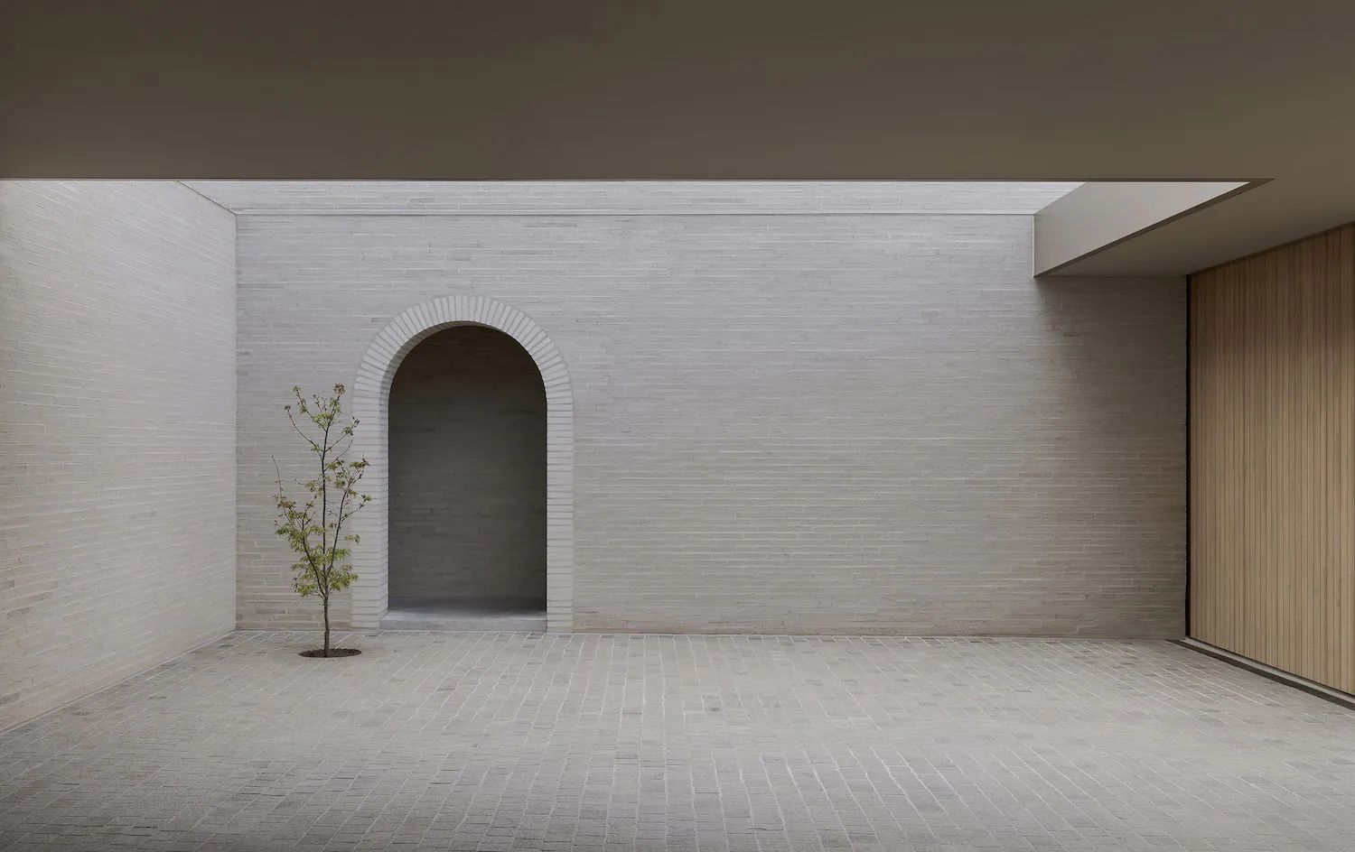 Minimalist interior courtyard with white brick walls, a central arched doorway, a small leafy tree, and a wooden slat wall on the right under an overcast sky.