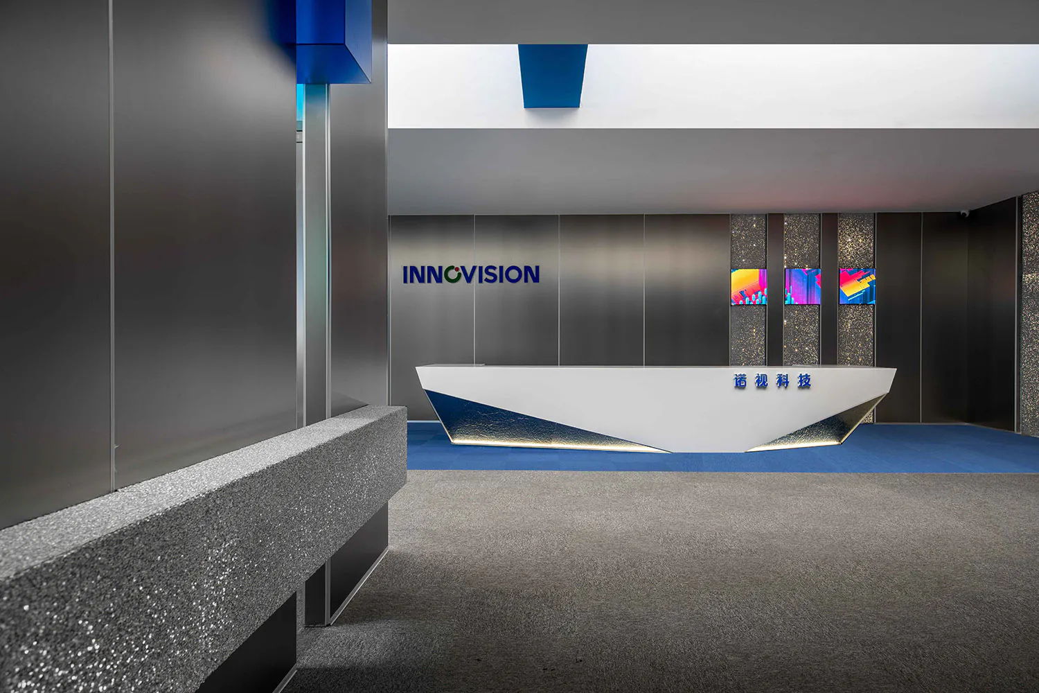 Modern corporate reception area featuring a white geometric reception desk with blue metallic base, stainless steel walls, blue carpet, and colorful digital art displays on the back wall with the company name INNOVISION.