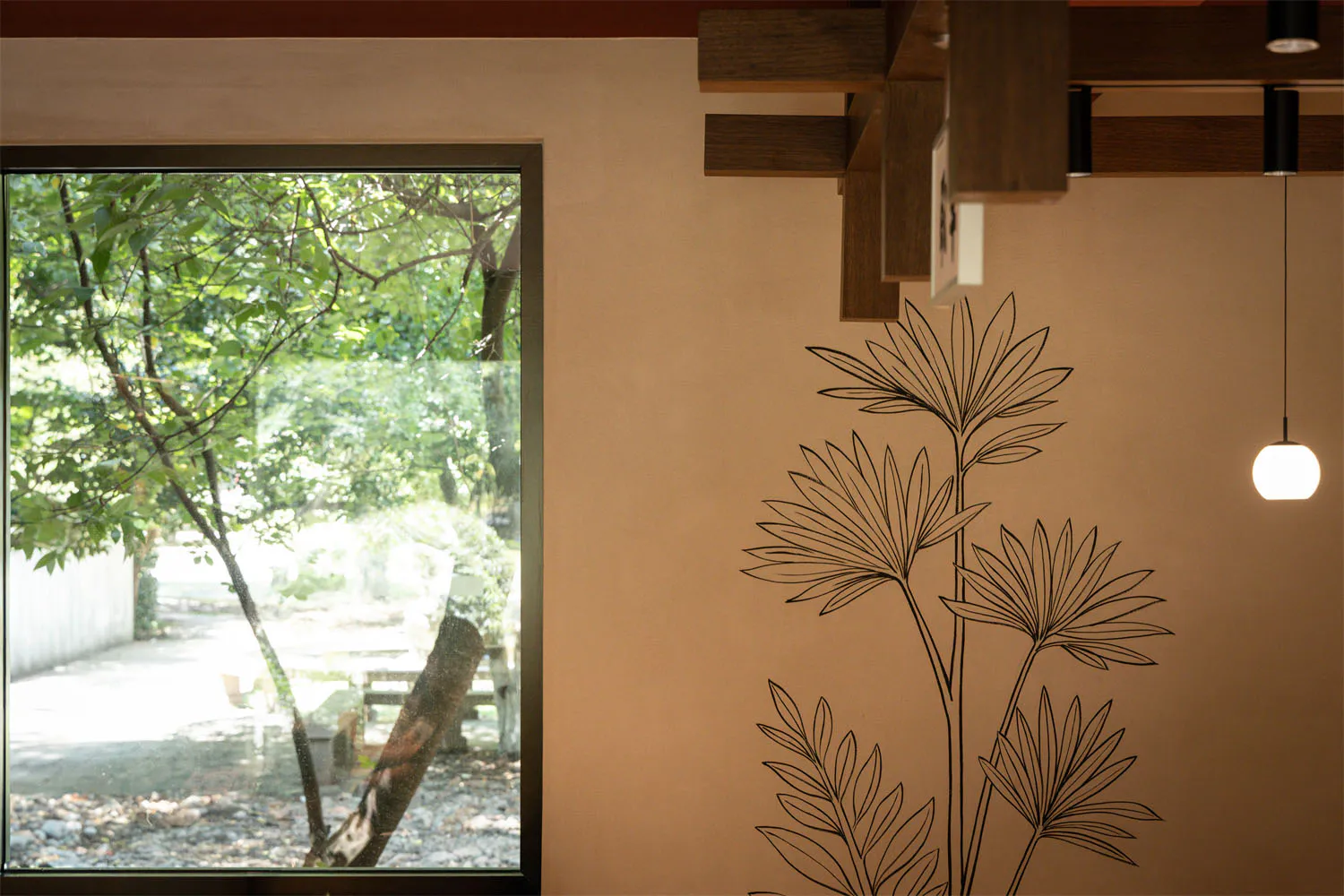 Interior corner featuring a large beige wall with black botanical line art, exposed wooden beams, a modern pendant light, and a large window overlooking a green garden.