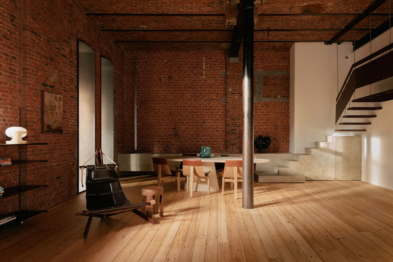 Spacious industrial loft interior featuring exposed red brick walls, a large round dining table with wooden chairs, a black leather sling chair, a concrete staircase, and dramatic natural light.