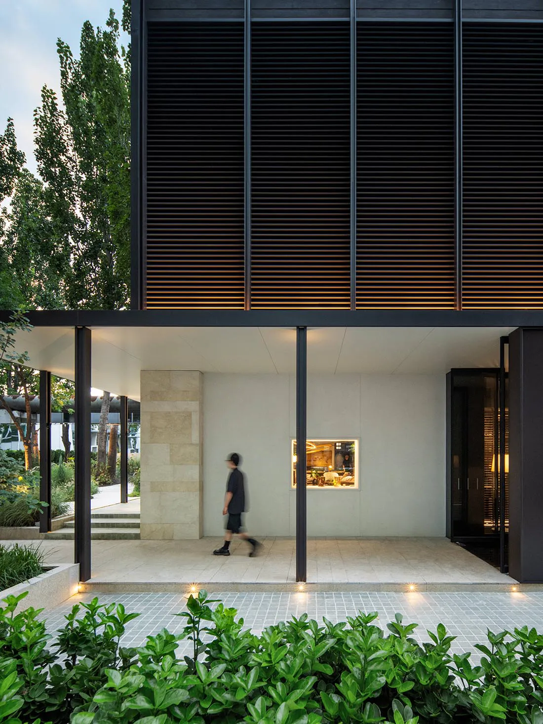 Modern minimalist courtyard of a contemporary house featuring a large travertine stone wall, dark horizontal wooden louvers with warm backlighting, a person walking across the paved area, and lush green foreground planting.