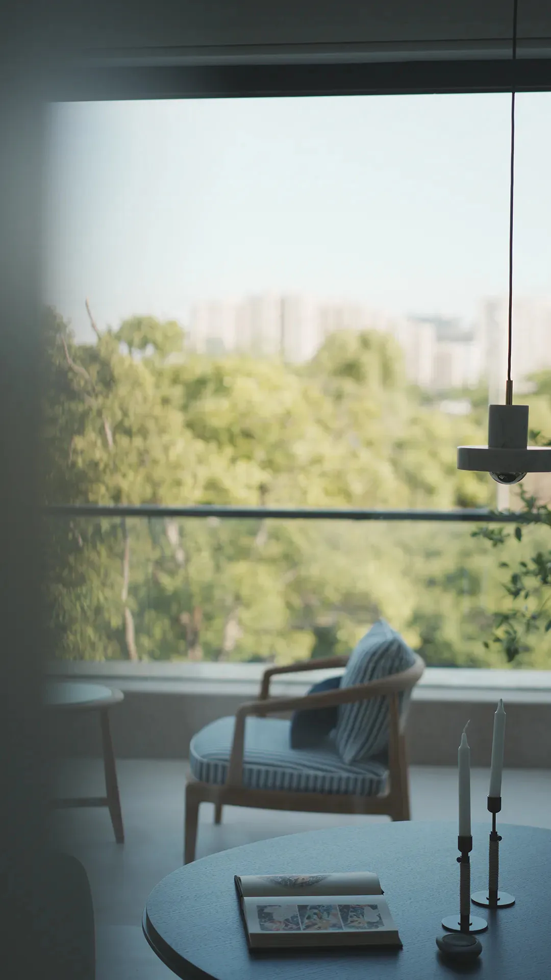 Modern wooden armchair with blue and white striped cushion positioned by a large window overlooking green trees and distant city buildings, with a round coffee table in the foreground holding an open book and two white taper candles.