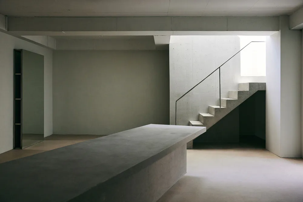 Empty minimalist brutalist interior featuring a raw concrete floating staircase, large concrete island, and soft natural light from a high window.